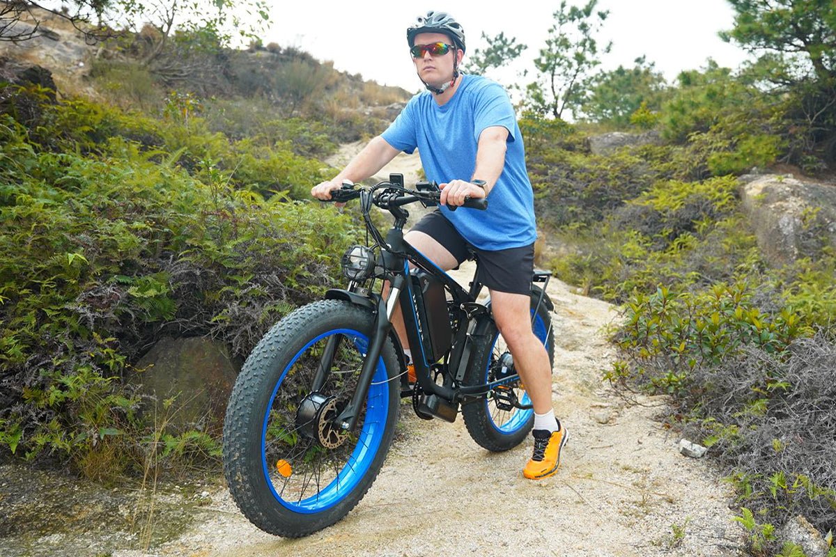 Helmeted rider seated on a fat-tire electric bike with blue wheel rims beside a rocky path